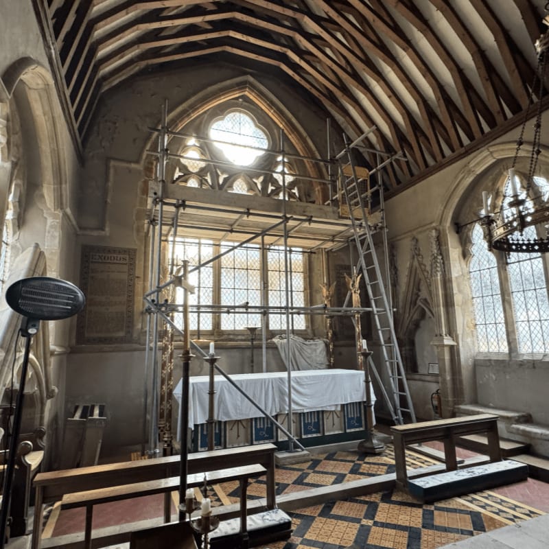 Scaffolding surrounds an altar in the foreground, while a large, ornate window and wooden ceiling are visible in the background.