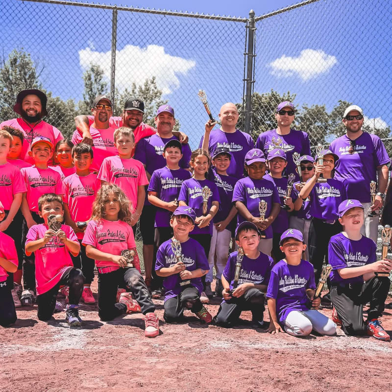 A group of children and adults in purple and pink shirts pose for a photo on a baseball field with a chain-link fence and trees in the background.