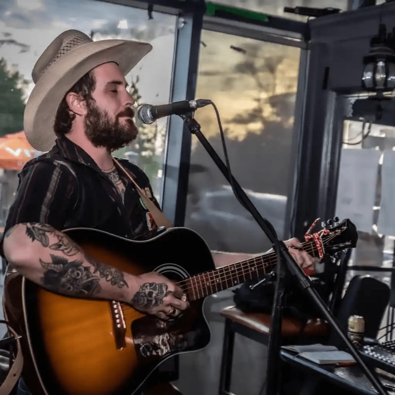 A man in a cowboy hat plays an acoustic guitar and sings into a microphone, with a blurred outdoor scene visible through a window in the background.