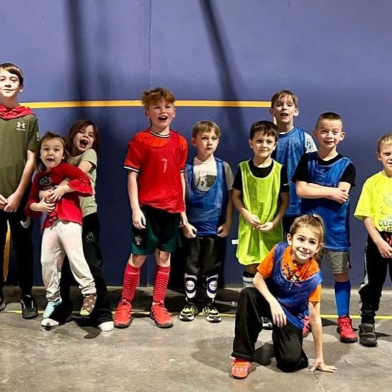 A group of children in athletic wear stand in front of a blue wall, with one child in the foreground on their knees.