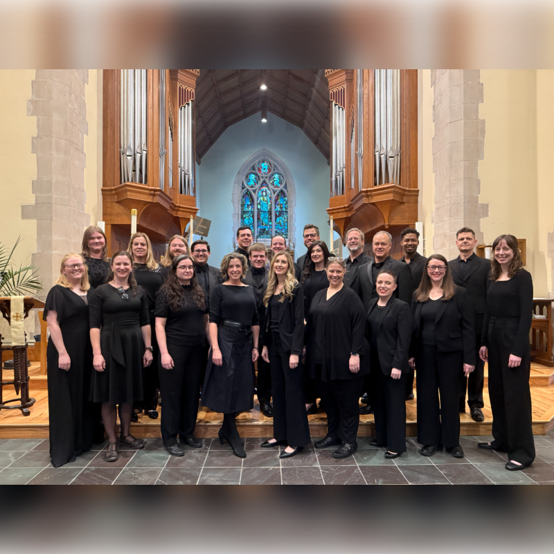 A choir stands in formal attire in front of a large pipe organ in a church.