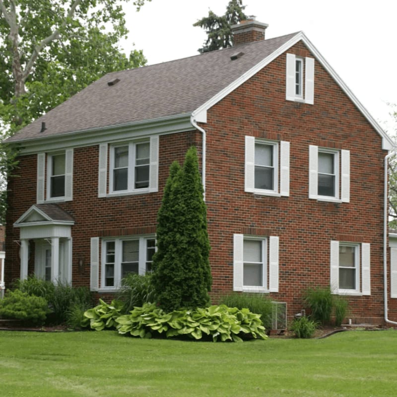 A red brick house with white shutters stands on a green lawn, with lush green bushes and a tall evergreen tree in front, and mature trees in the background.