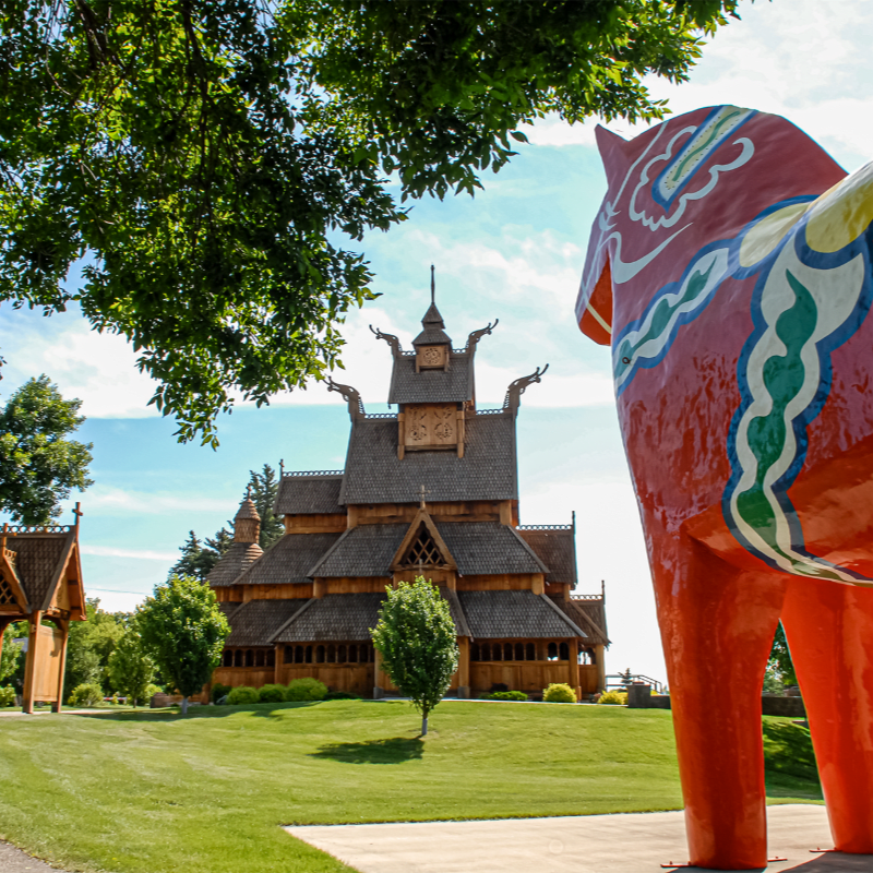A large, brightly painted red horse statue stands in the foreground, with a wooden church and green trees in the background under a blue sky.