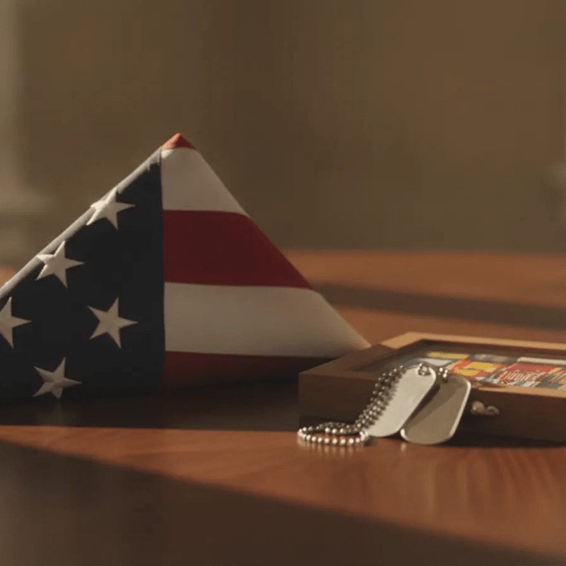 A folded American flag and dog tags rest on a wooden surface in front of a blurred background.
