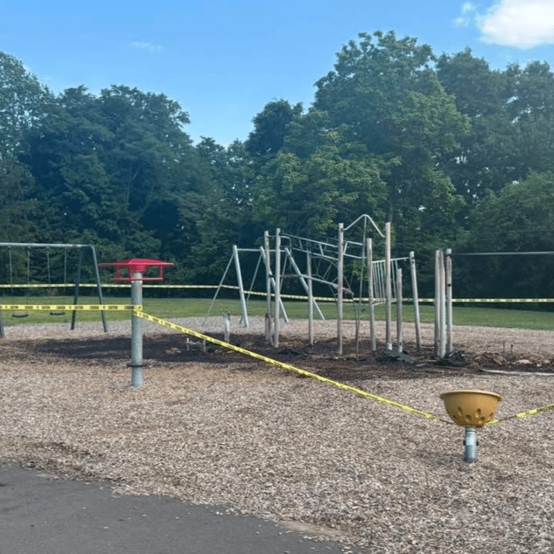 Yellow caution tape surrounds a damaged playground with swings and climbing structures, set against a backdrop of lush green trees under a cloudy sky.