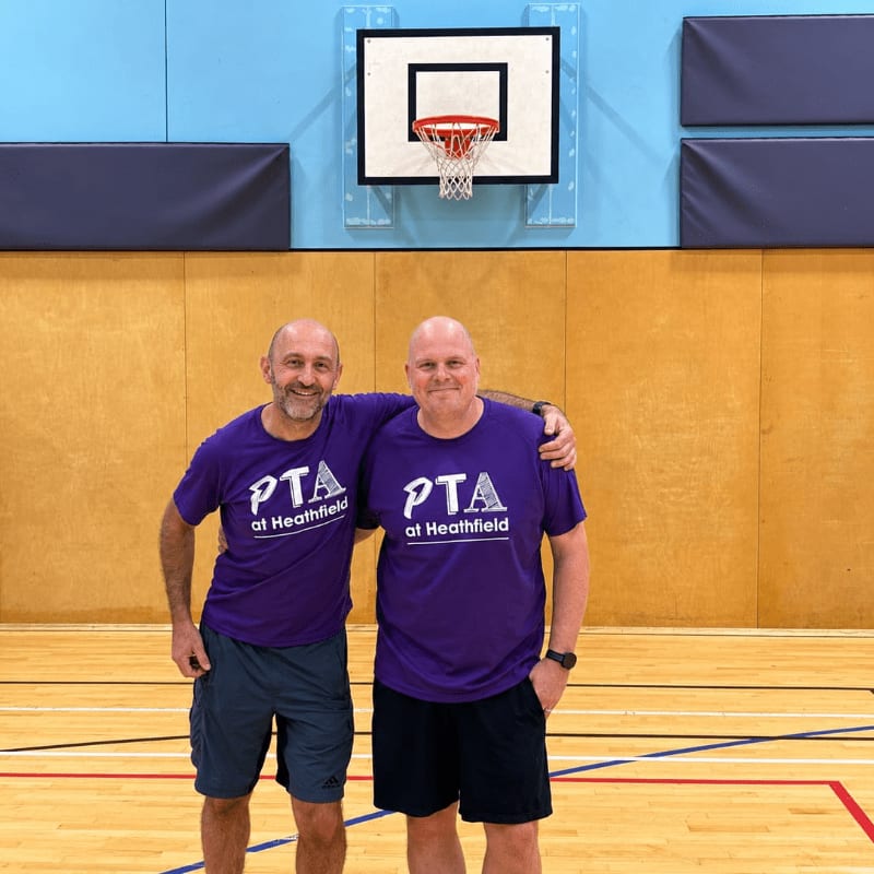 Two men in purple "PTA at Heathfield" t-shirts stand with arms around each other in a gymnasium with a basketball hoop in the background.
