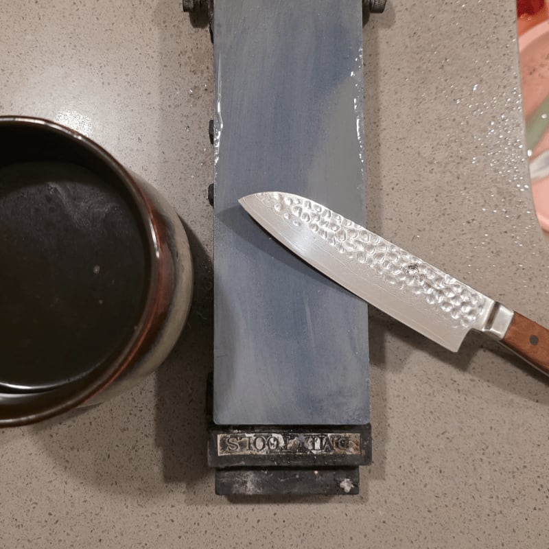 A hammered metal knife rests on a wet sharpening stone, with a small bowl of dark liquid to its left and a blurred pink object in the background.