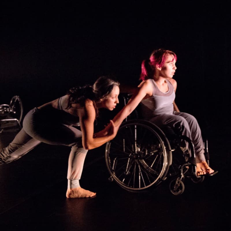 A dancer in the foreground supports a person in a wheelchair against a dark background.