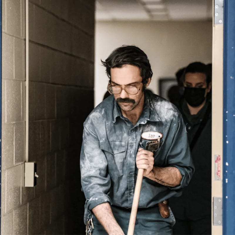 A man in a dusty blue uniform with a mustache and glasses sweeps the floor in the foreground, while another person wearing a mask stands in the blurred background.
