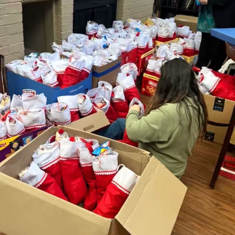 A woman in the foreground is surrounded by numerous Christmas stockings filled with gifts, while in the background, more stockings and boxes are stacked high.