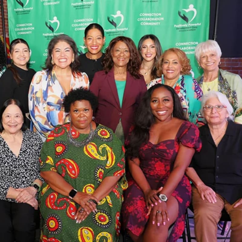 A diverse group of ten women pose for a photo in front of a green backdrop with a logo, with some women seated in the foreground and others standing behind them.