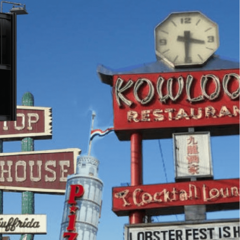 A bright blue sky serves as the backdrop for a collection of colorful neon signs, including "Kowloon Restaurant" and "Cocktail Lounge," with a tall, cylindrical building and other signs in the foreground.