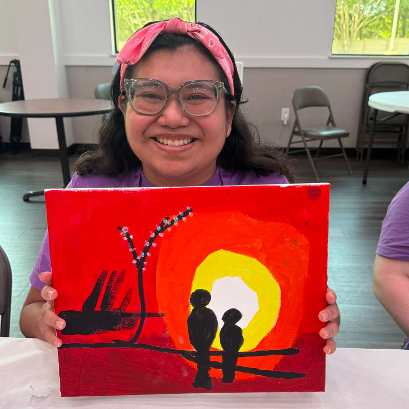 A smiling young woman in the foreground holds up a vibrant sunset painting, while the background shows a room with tables and chairs.