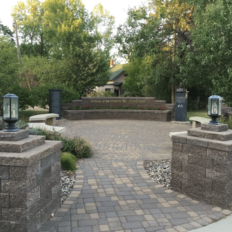 A paved walkway leads to a memorial plaza with stone pillars and lanterns in the foreground, while a building and lush trees form the background.