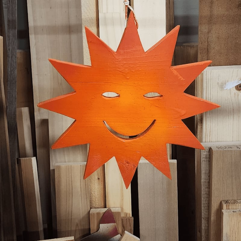 A bright orange, smiling sun decoration hangs in front of a background of stacked wooden planks.