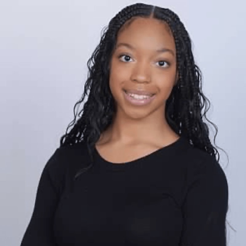 A young Black woman with braided hair smiles at the camera in front of a plain gray background.
