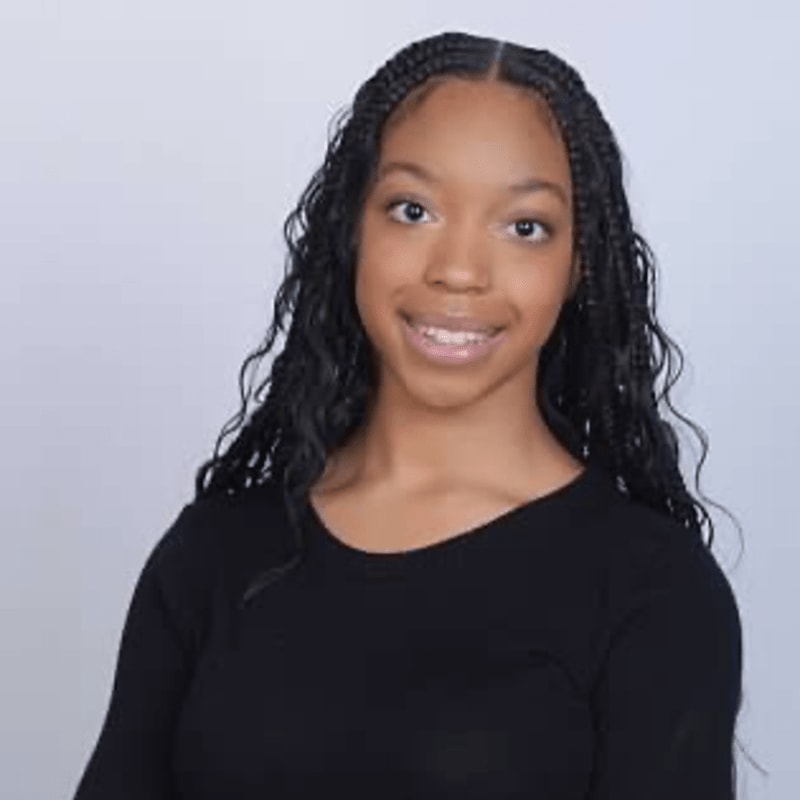 A young Black woman with braided hair smiles against a plain, light gray background.