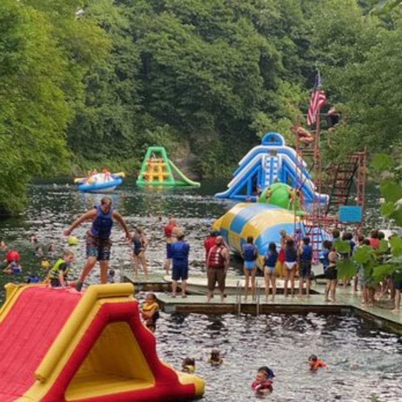 People enjoy inflatable water slides and obstacles in a lake surrounded by lush green trees.