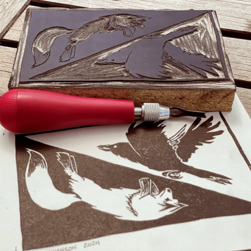 A linocut block with a carved fox and bird design rests above a freshly printed impression of the same design, with a carving tool in the foreground.