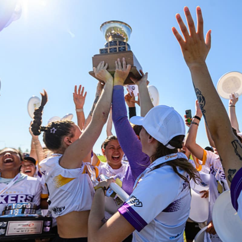 A group of jubilant women in purple and white uniforms raise a championship trophy against a bright blue sky.