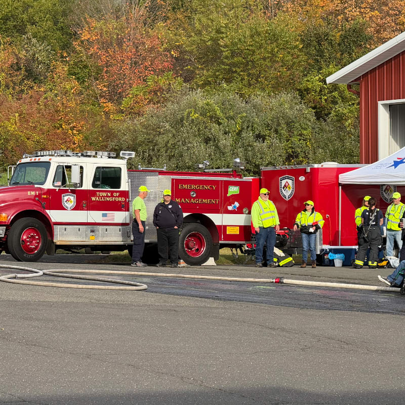 A fire truck and emergency management personnel stand in front of a red building with autumn foliage in the background.