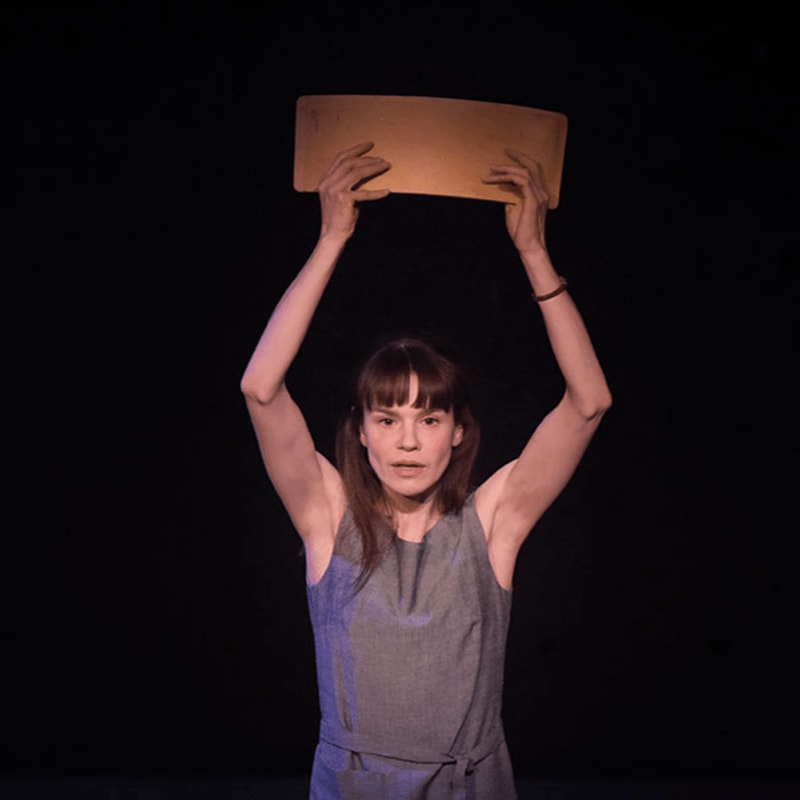 A woman in a gray dress holds a rectangular object above her head against a dark background.