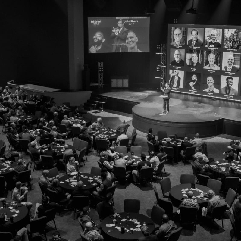 A speaker stands on a stage in front of a large screen displaying portraits, while an audience sits at round tables in the foreground.