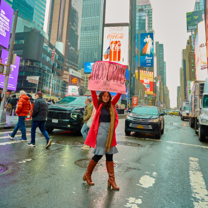 A woman holds a sign in the foreground of a bustling, wet Times Square, with towering billboards and traffic filling the background.