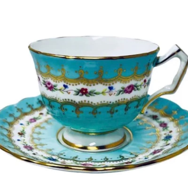 A turquoise teacup and saucer with gold trim and floral accents sits against a white background.