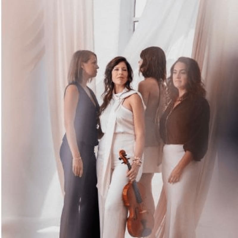 Four women in elegant attire stand in a softly lit room with sheer drapes in the background, one woman in the foreground holding a violin.