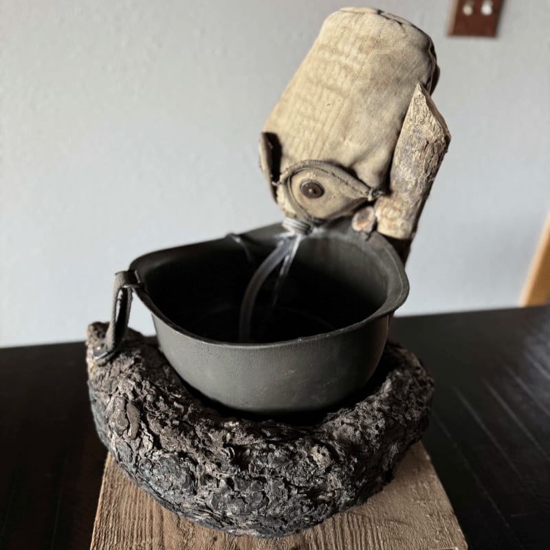 A rustic fountain made from a helmet and rough materials sits on a wooden surface against a plain wall.