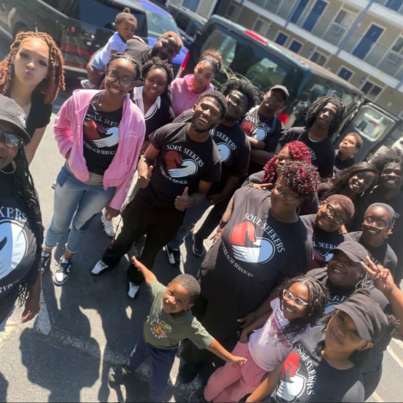 A group of people, many wearing matching t-shirts, pose for a photo in front of a building and vehicles.