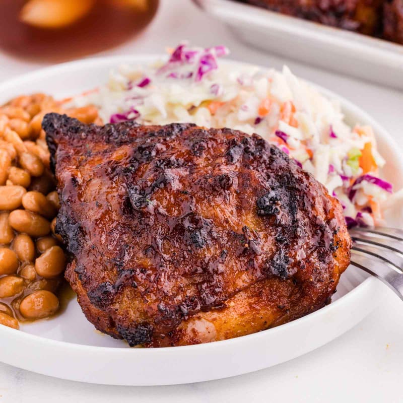 A glistening piece of barbecued chicken sits on a white plate with baked beans and coleslaw, while a glass of iced tea is visible in the blurred background.