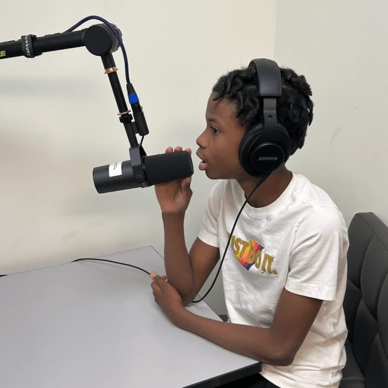 A young boy wearing headphones speaks into a microphone at a desk, with a plain wall in the background.