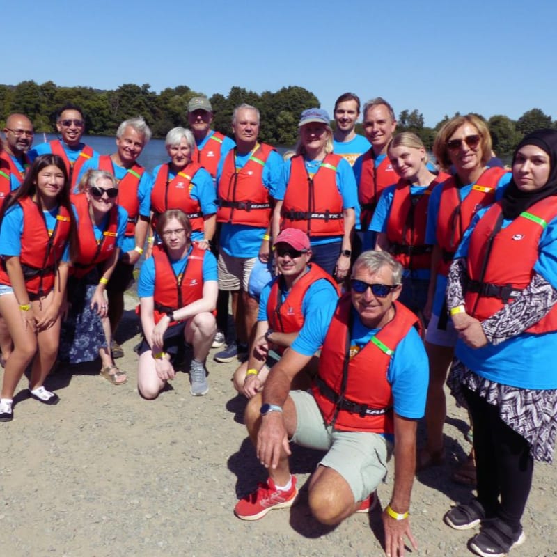 A group of people in life vests stand on a sandy shore with trees and a clear blue sky in the background.