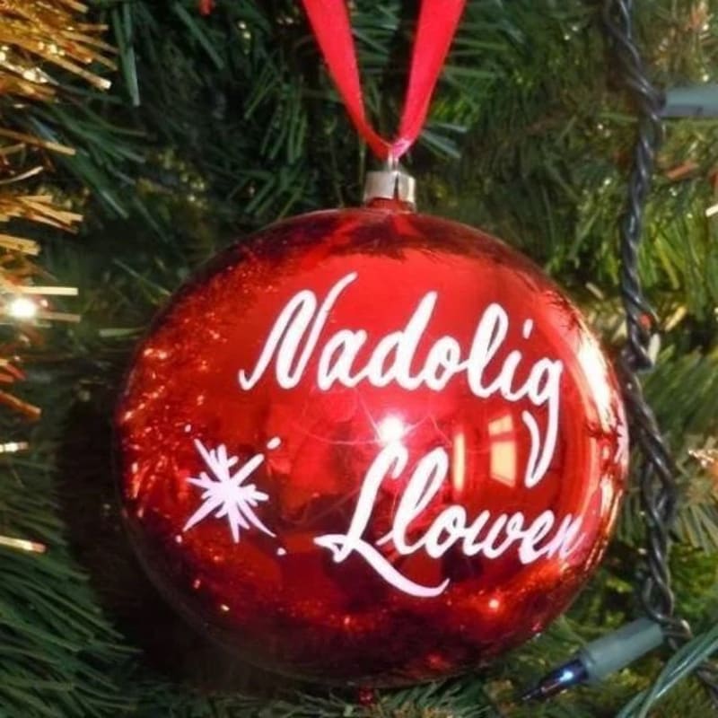 A shiny red Christmas ornament with "Nadolig Llawen" written in white script hangs from a decorated Christmas tree with twinkling lights in the background.