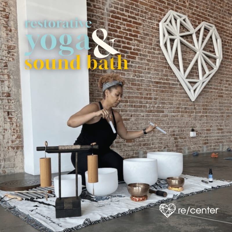 A woman in the foreground plays crystal singing bowls and chimes, while a large geometric heart adorns a brick wall in the background.