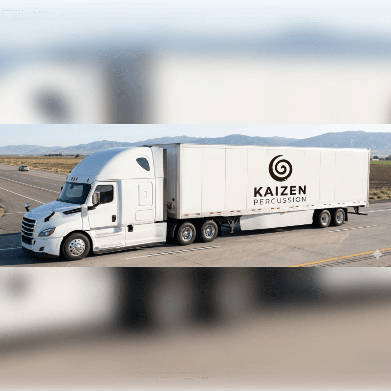 A white semi-truck with "KAIZEN PERCUSSION" on its trailer drives on a highway with mountains in the background.