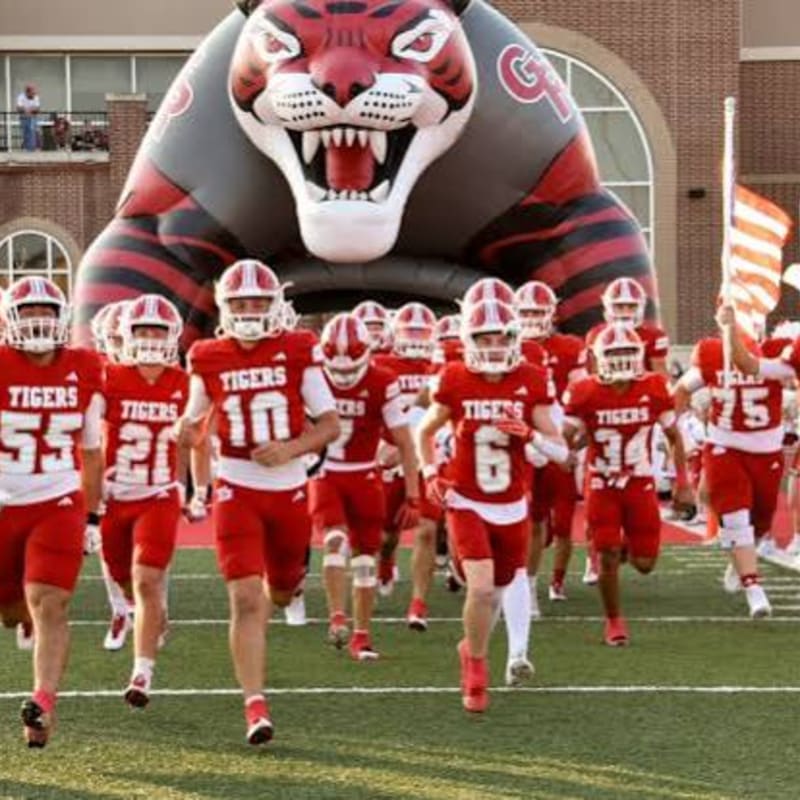 A football team in red and white uniforms runs onto the field through a large inflatable tiger head tunnel.
