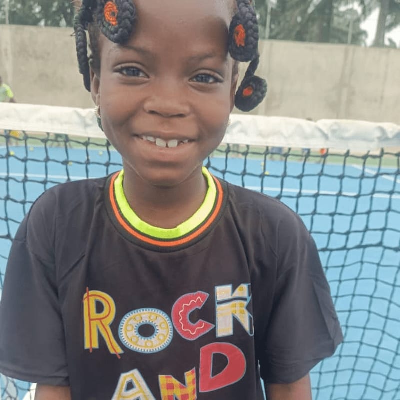 A young girl with braided hair smiles at the camera in the foreground, with a blue tennis net and court visible in the background.