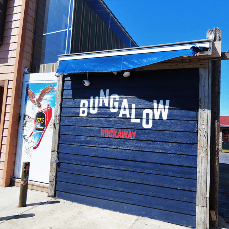 A blue wooden facade with "BUNGALOW ROCKAWAY" painted in white and red letters stands in the foreground, while a building with a striped awning and a white door with an eagle graphic is visible to the left, against a clear blue sky.