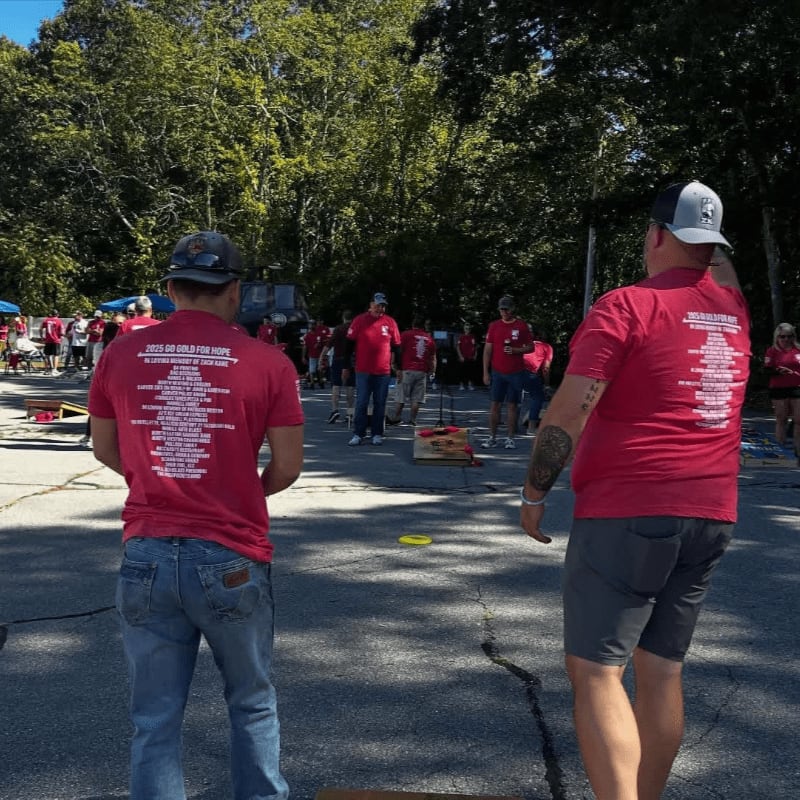 Two men in red t-shirts play cornhole on a paved surface with trees and a crowd in the background.