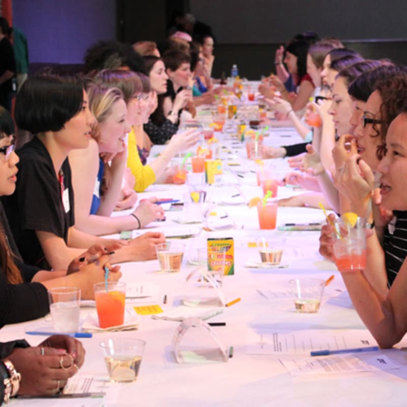 A long table filled with women enjoying drinks and conversation is set against a blurred background of more attendees.