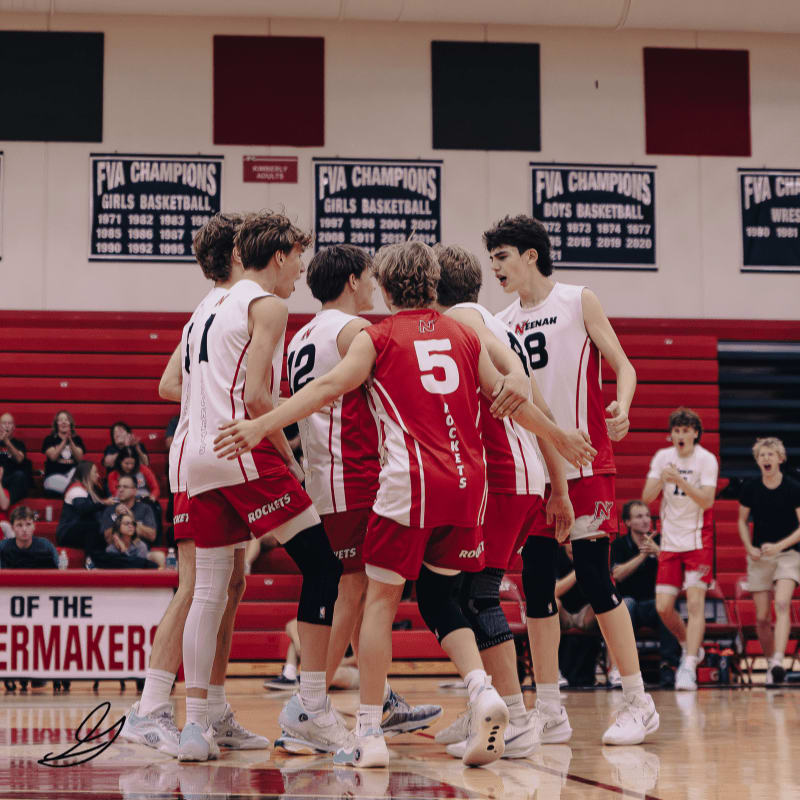 A group of young male volleyball players in red and white uniforms huddle together on a wooden court, with banners celebrating past championships hanging in the background.