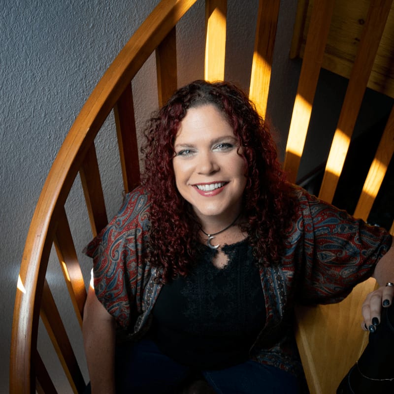 A smiling woman with curly red hair sits on a wooden spiral staircase, with sunlight casting shadows in the background.