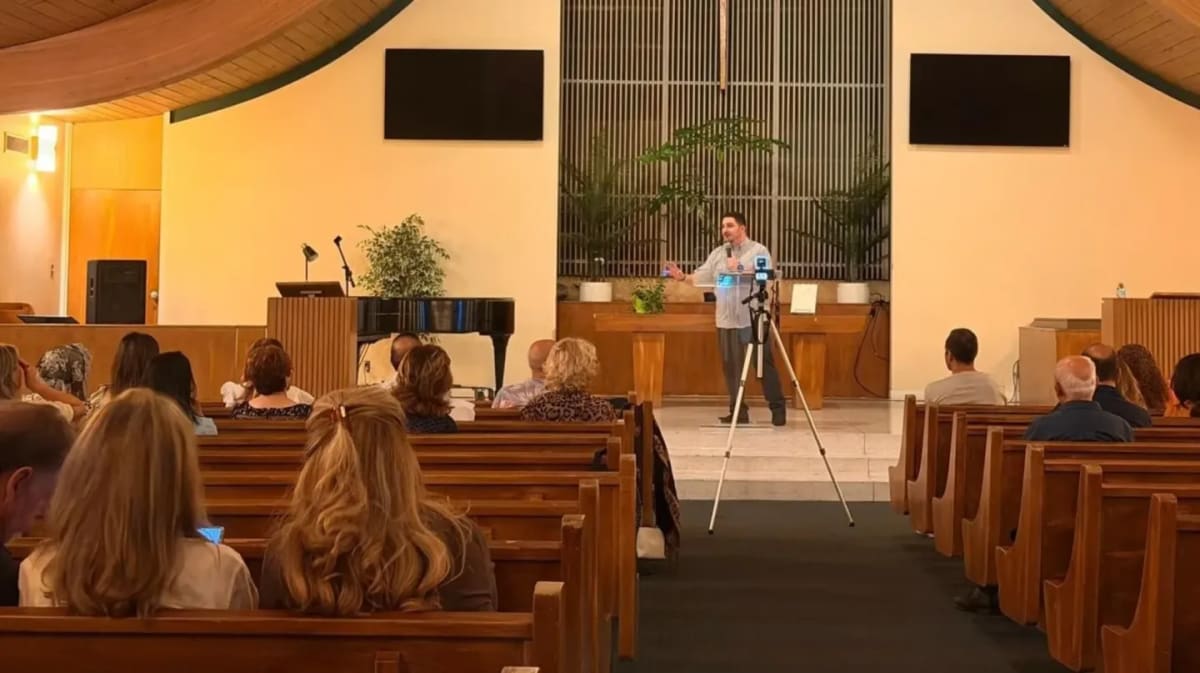 A man stands at a podium in front of a congregation seated in pews, with screens mounted on the wall behind him.