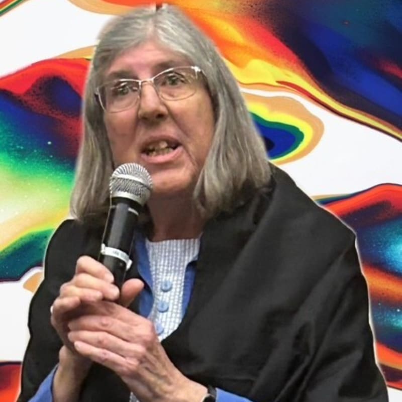 A woman with gray hair and glasses speaks into a microphone, with a vibrant, abstract painting behind her.
