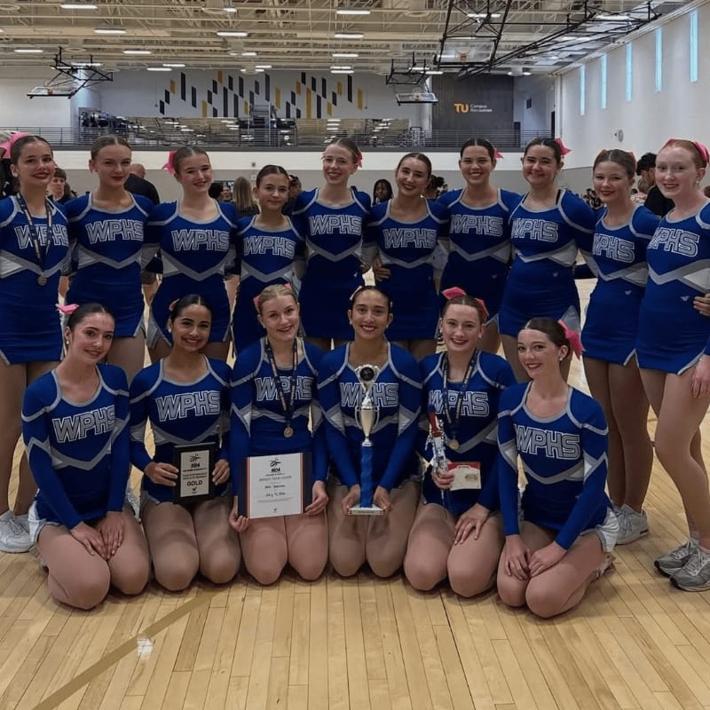 A group of young women in blue and white cheerleading uniforms pose with trophies in the foreground, while bleachers and a gymnasium are visible in the background.