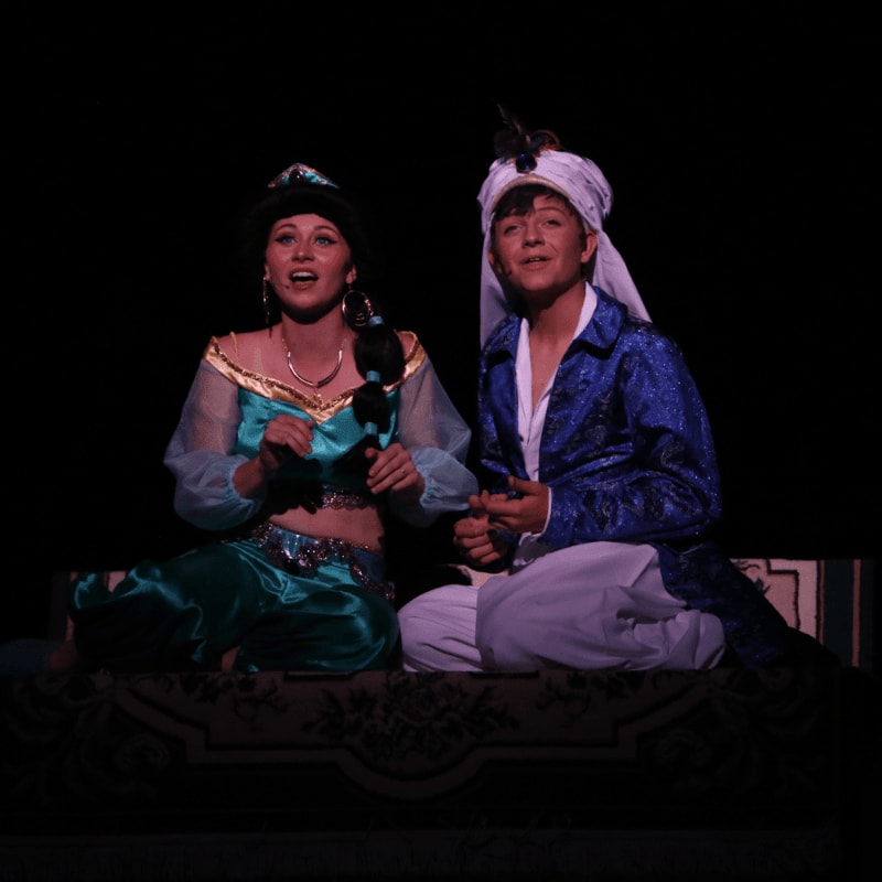 A young woman in a teal costume and a young man in a blue and white outfit sit on a patterned rug against a dark background.