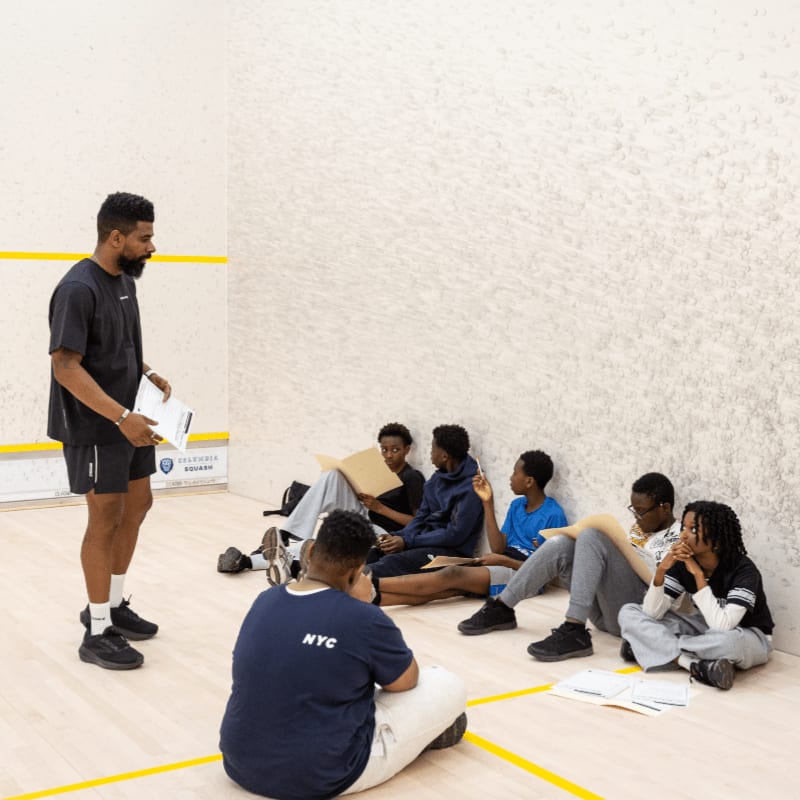 A man stands addressing a group of young people sitting on the floor of a squash court.
